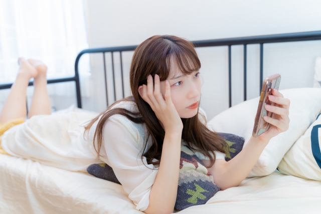 布団にうつ伏せで寝そべりながら不機嫌そうな顔でスマホを見つめる女性