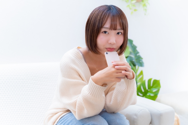 白いソファに座り、スマホを両手で持ちながら真顔でこちらを見つめてくる女性