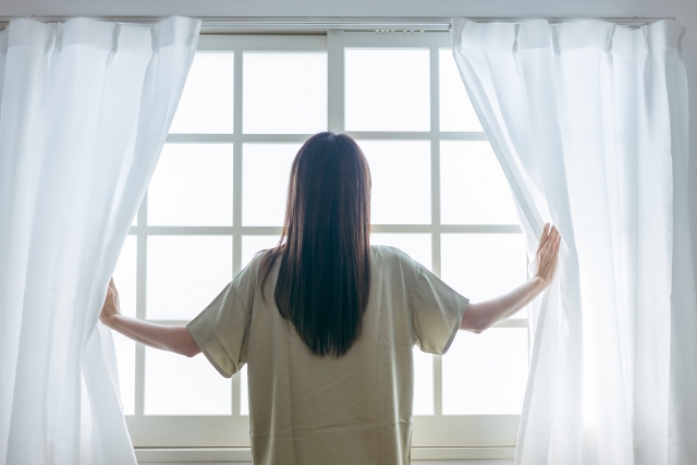 窓のカーテンを上げて朝の空を見上げている女性の後ろ姿