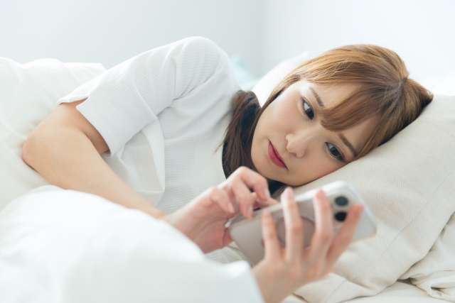 ベッドに左半身を下にして横たわりスマホをいじる女性