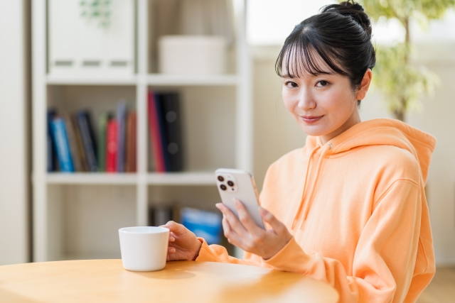 椅子に座り、片手にコーヒーカップを持ち、もう片方の手でスマホを持ち、カメラ目線でこちらを見つめているオレンジ色のパーカーを着た笑顔の女性