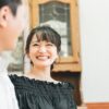 屋内で会話をするカップル。男性は向こう側を向いておりよく分からないが、女性はこちら向きで、男性に満面の笑みを浮かべている。