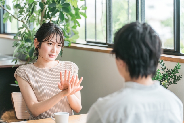 カフェで向かい合って座っている男女。男性は向こう側を向いている。女性はこちら側を向いて男性の正面に座っており、体の前で手首をクロスさせてバッテンを作ることで、男性に向けて何かを拒否する意思を表現している。