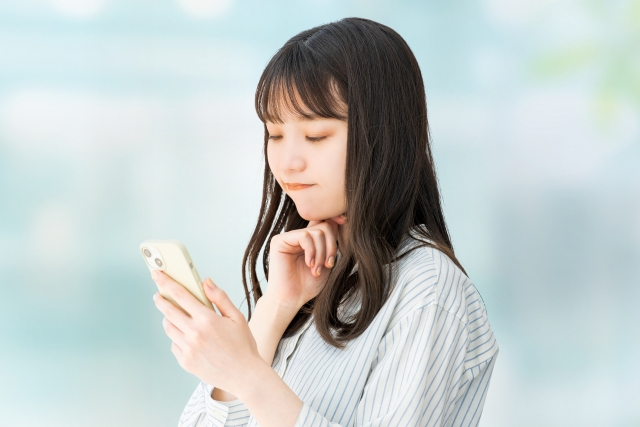 顎に手を添えた若い女性がスマートフォンを持ちながら下を見て、考え込んでいるような表情をしている。