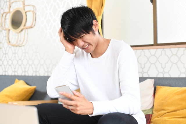 室内にて白い服の男性がスマートフォンを見ながら悩んでいる