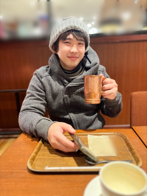 冬のカフェでニット帽とグレーのパーカーを着た若い男性が、笑顔でコップを手にしている。