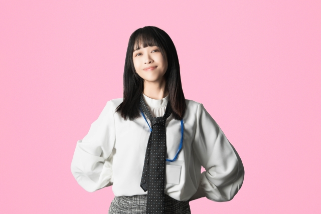 白い服にネクタイ姿の若い女性が、腰に手を当てて自信ありげに笑っている。