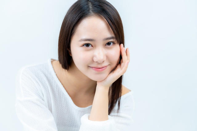 白いトップスを着た若い女性が、手を頬に添えてやさしく微笑んでいるバストアップ写真。