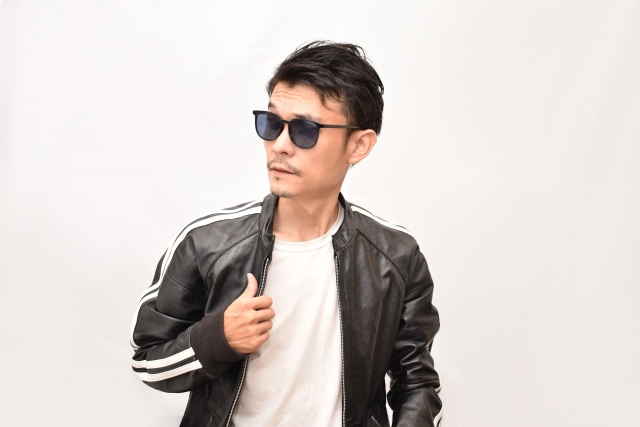 白い背景の前で革ジャンを着てサングラスをかけた男性が、クールなポーズでキメている。