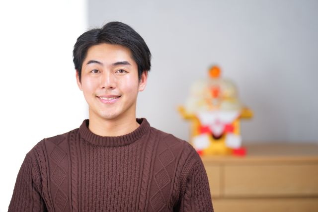 茶色いセーターを着た男性が、正面を向いて穏やかに微笑んでいる室内ポートレート。