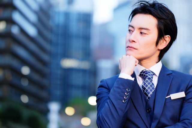スーツ姿の男性が都会のビル街で腕を組み、真剣な表情で何かを考えている